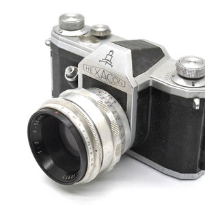 Può includere: Una macchina fotografica a pellicola da 35 mm argentata e nera con un obiettivo Hexacon. La macchina fotografica ha un rivestimento in pelle nera e un corpo in metallo argentato. L'obiettivo è un obiettivo 1:2 F=58 mm.
