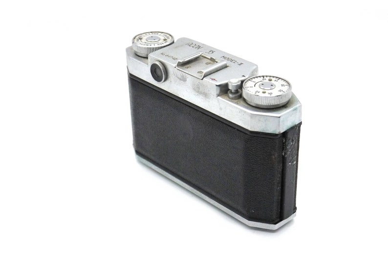 Pu&ograve; includere: Una macchina fotografica a pellicola da 35 mm argentata e nera con un rivestimento in pelle nera. La macchina fotografica ha un obiettivo argentato e un quadrante argentato sul lato destro. La macchina fotografica ha il testo "ALPA" e "Model-1" sulla parte superiore.