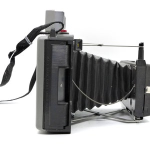 May include: A gray and black vintage Polaroid camera with a black strap and a bellows lens. The camera has a red button and a black viewfinder.