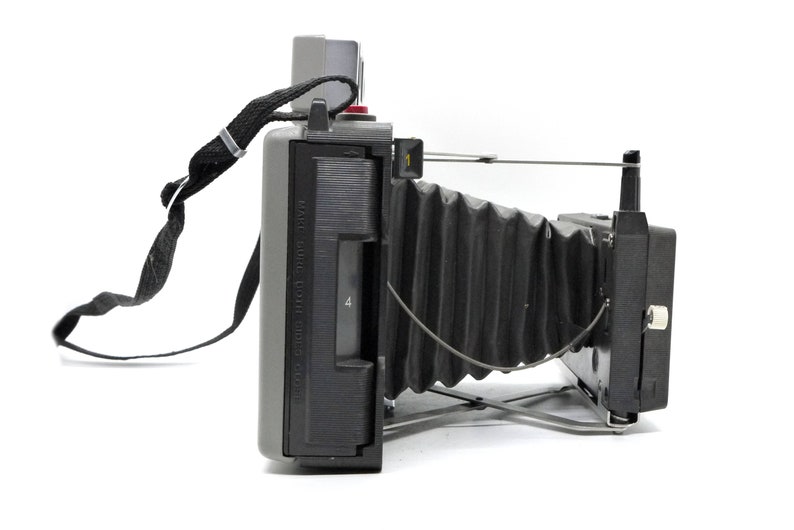 May include: A gray and black vintage Polaroid camera with a black strap and a bellows lens. The camera has a red button and a black viewfinder.