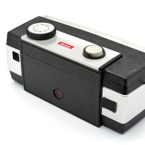 May include: A black and silver Kodak Instamatic camera with a red viewfinder. The camera has a wind knob with the word "WIND" printed on it.