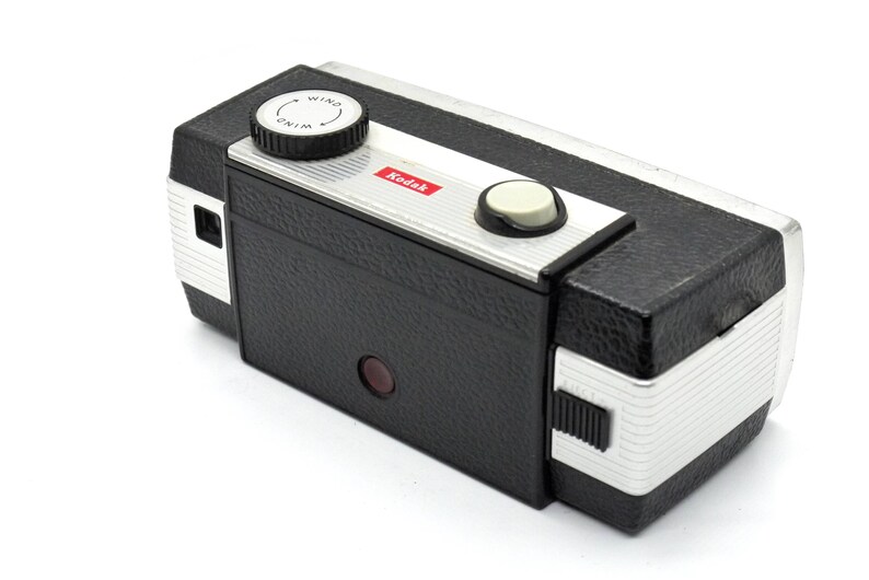 May include: A black and silver Kodak Instamatic camera with a red viewfinder. The camera has a wind knob with the word "WIND" printed on it.