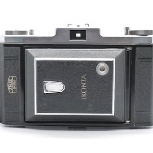 Puede incluir: Una c&aacute;mara Zeiss Ikonta negra y plateada con una correa de cuero. La c&aacute;mara tiene un visor y una tapa de lente.