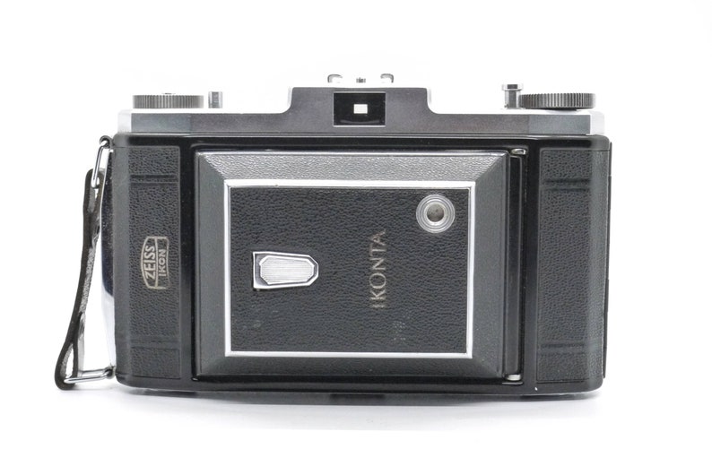 Puede incluir: Una c&aacute;mara Zeiss Ikonta negra y plateada con una correa de cuero. La c&aacute;mara tiene un visor y una tapa de lente.