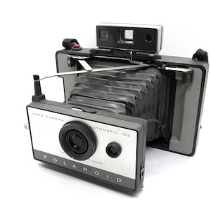 May include: A vintage Polaroid Land Camera Automatic 103 in black and silver with a bellows lens and a viewfinder. The camera has a strap attached.