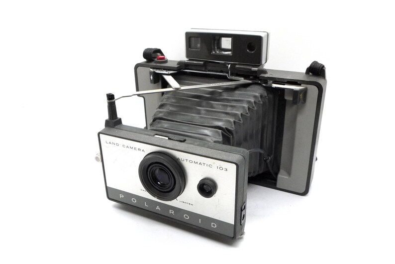 May include: A vintage Polaroid Land Camera Automatic 103 in black and silver with a bellows lens and a viewfinder. The camera has a strap attached.