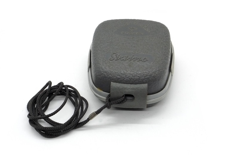 Puede incluir: Una caja rectangular gris texturizada con la palabra "Sixims" impresa en ella. La caja tiene un cord&oacute;n negro adjunto.