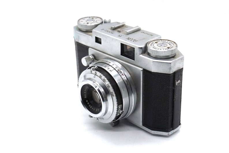 Pu&ograve; includere: Una macchina fotografica a pellicola da 35 mm di colore argento e nero con un rivestimento in pelle nera. La macchina fotografica ha un obiettivo con un anello argentato e un tappo dell'obiettivo nero. La macchina fotografica ha una piastra superiore argentata con il testo "RODA" e "F=2.8/50mm" stampato su di essa.