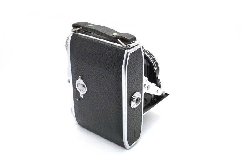 May include: A vintage folding camera with a black leather covering and a carrying handle. The camera is made of silver and black.