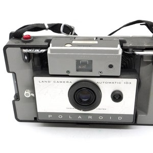 May include: A vintage Polaroid Land Camera Automatic 103 in gray and white with a black strap. The camera has a lens, a viewfinder, and a flash.