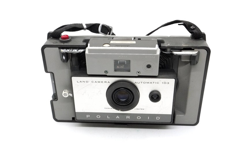 May include: A vintage Polaroid Land Camera Automatic 103 in gray and white with a black strap. The camera has a lens, a viewfinder, and a flash.