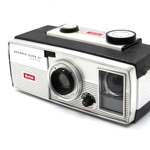 May include: A silver and black Kodak Brownie Super 27 camera with a red "Kodak" logo and a black "Brownie Super 27 Camera" label. The camera has a black lens with a silver ring and a black wind knob.