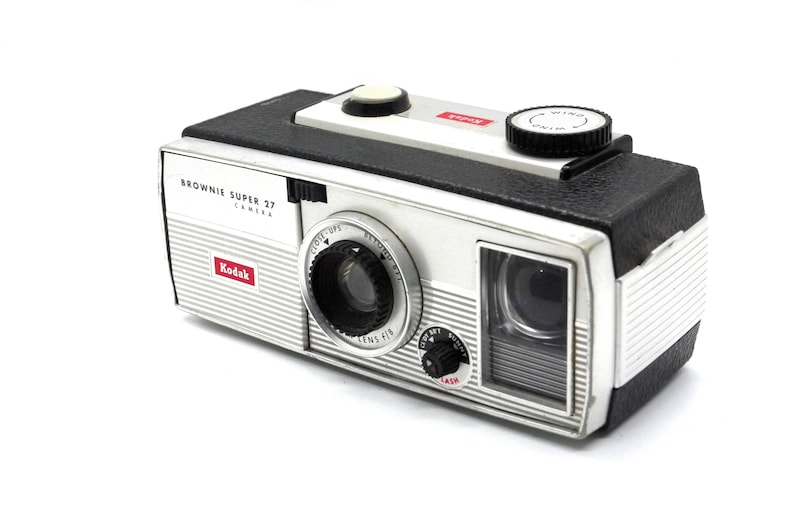 May include: A silver and black Kodak Brownie Super 27 camera with a red "Kodak" logo and a black "Brownie Super 27 Camera" label. The camera has a black lens with a silver ring and a black wind knob.