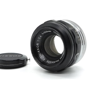 May include: A black Nikon 105mm f/1.5 E-Nikkor lens with a silver aperture ring. The lens has a black lens cap with the word "Nikkor" printed in white.