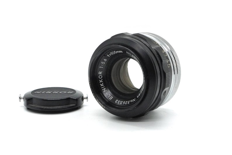May include: A black Nikon 105mm f/1.5 E-Nikkor lens with a silver aperture ring. The lens has a black lens cap with the word "Nikkor" printed in white.