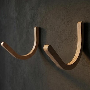 May include: Two J-shaped wall hooks crafted from light-coloured wood with visible grain patterns. Each hook features a curved, smooth design with a rounded tip, mounted securely to provide a functional storage solution for hanging various items.