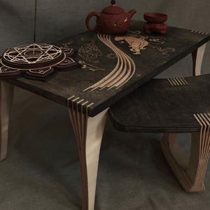 May include: A dark wood nesting table set featuring light-colored layered plywood legs and intricate laser-cut inlays of a tiger and floral patterns on the surface. A circular lotus-shaped incense holder and a small red clay teapot sit on top.