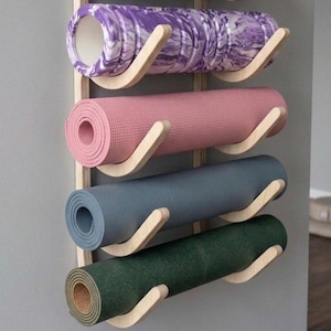 May include: A wall-mounted wooden rack featuring four curved supports holding four rolled exercise mats. The mats are colored purple with a white marble pattern, solid light pink, solid slate blue, and solid dark forest green with a cork base.