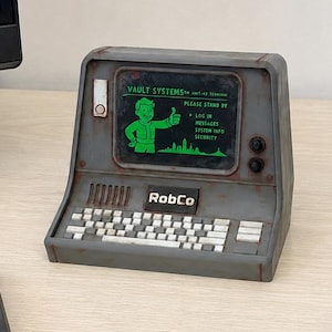 May include: A grey, weathered-look desktop terminal replica featuring a screen with green text reading VAULT SYSTEMS, PLEASE STAND BY, LOG IN, MESSAGES, SYSTEM INFO, SECURITY, and a Vault Boy graphic. The unit includes a keyboard and RobCo label.