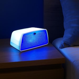 Può includere: Una lampada da comodino bianca e blu con un pannello frontale perforato, che emette una luce blu brillante. La lampada è rettangolare con bordi arrotondati e si trova su un comodino di legno accanto a un letto con un cuscino grigio.