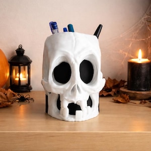 May include: A white skull-shaped pen holder with black eye sockets and mouth. The Halloween-themed desk accessory holds pens and is surrounded by a carved pumpkin, a lit candle, and a small skull on a wooden surface.