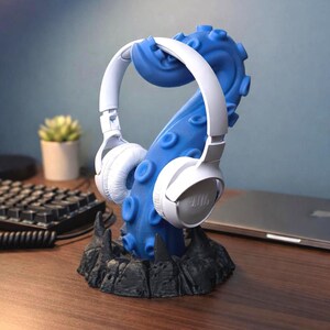 Può includere: Un supporto per cuffie a forma di tentacolo di polpo blu con cuffie bianche. Il supporto ha una base nera. Le cuffie sono bianche con il marchio "JBL" visibile. Il supporto è stampato in 3D.