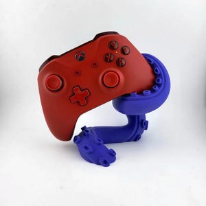 Puede incluir: Un mando de Xbox rojo está sujeto por un soporte azul en forma de pulpo. El mando tiene botones rojos y una cruz direccional roja.