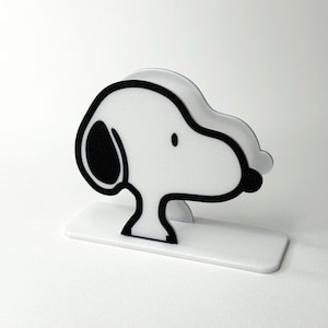 Może przedstawiać: Biało-czarna figurka Snoopy'ego, psa z kreskówki, stoi na prostokątnej podstawie. Figurka ma czarny kontur i czarne detale uszu i nosa. Prosty i nowoczesny design, odpowiedni do dekoracji wnętrz.