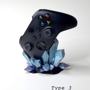 May include: A black Xbox controller is displayed on a crystal-shaped stand. The stand is made of blue and purple plastic and resembles a cluster of crystals. The text "Type 3" is printed below the stand.