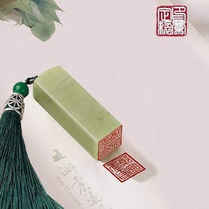 May include: A rectangular, pale green jade seal with red Chinese characters stamped on the bottom. A matching red stamp is next to the seal. A dark green tassel with a silver-colored bead hangs from the seal.
