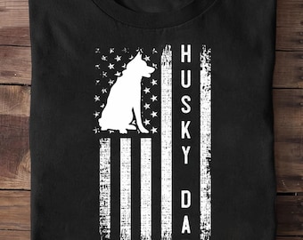 May include: Black t-shirt featuring a white silhouette of a husky dog within a distressed American flag design. The words "HUSKY DAD" are printed vertically in white on the right side of the flag. The shirt is folded.