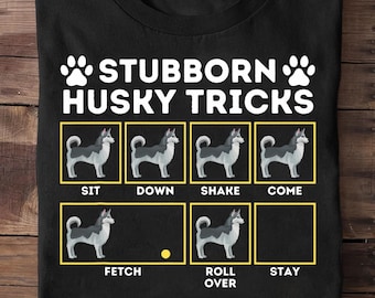 May include: Black t-shirt with the text "STUBBORN HUSKY TRICKS" in white. The shirt features illustrations of a husky dog demonstrating commands like sit, down, shake, come, fetch, roll over, and stay, each in a yellow box.