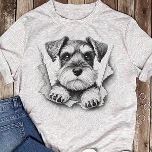 May include: A light grey t-shirt with a detailed black and white illustration of a Schnauzer dog. The dog is depicted as breaking through a torn section of the shirt, with its paws visible.