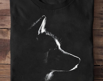 May include: Black t-shirt featuring a striking white silhouette of a dog's profile. The design highlights the dog's ear, snout, and fur with a minimalist, artistic style. The shirt is folded on a wooden surface.