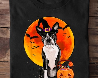 Puede incluir: Camiseta negra con un Boston Terrier con un sombrero de bruja con hebilla morada, sobre una gran luna naranja. Murciélagos volando alrededor de la luna. Una cesta de calabaza llena de caramelos está a los pies del perro. Temática de Halloween.