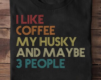 May include: Black t-shirt with the text "I LIKE COFFEE MY HUSKY AND MAYBE 3 PEOPLE" in retro-style lettering. The text is in red, orange, yellow, and teal. The shirt is folded on a wooden surface.