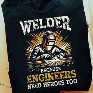 May include: Black t-shirt with a graphic of a welder wearing a welding mask and holding a welding torch. The text "Welder because engineers need heroes too" is printed on the shirt.