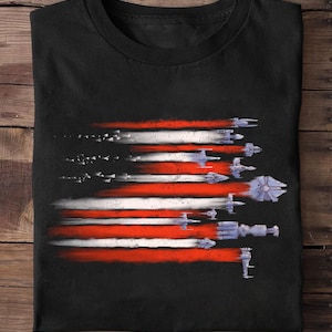 May include: Black t-shirt featuring a graphic design of the American flag with red and white stripes. The stripes are formed by a series of spaceships, with a blue and gray color scheme. The design is a unique take on a patriotic theme.