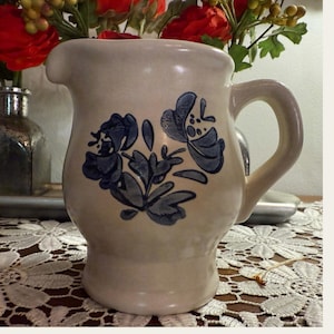 May include: A small, cream-colored ceramic pitcher with a blue floral design. The pitcher has a rounded body and a handle.