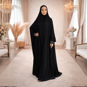 Peut inclure: Une femme porte un abaya noir avec des poignets ornés et un hijab assorti. Elle se tient dans une pièce bien éclairée. L'abaya est longue et fluide. Le fond comprend des meubles et une décoration élégants.