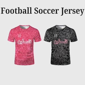 May include: Two short-sleeved football jerseys. One is a vibrant pink with a floral pattern, the other is black with a similar floral design. Both jerseys feature the word "Palestine" in Arabic script.