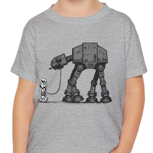 May include: A gray t-shirt featuring a black and white illustration of a Stormtrooper walking an AT-AT walker on a leash. The design is a humorous take on a classic science fiction theme.