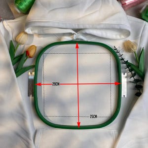 May include: A white hooded sweatshirt with a green embroidery hoop. The hoop measures 29cm by 25cm. The sweatshirt has a front pocket and a drawstring hood. The background includes spools of thread and artificial flowers.