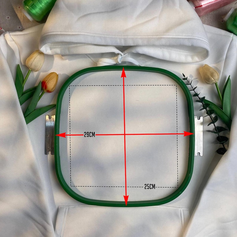 May include: A white hooded sweatshirt with a green embroidery hoop. The hoop measures 29cm by 25cm. The sweatshirt has a front pocket and a drawstring hood. The background includes spools of thread and artificial flowers.