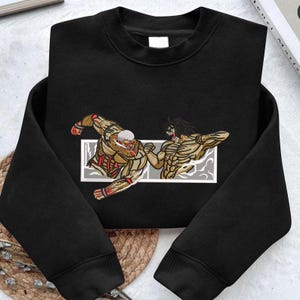 May include: Black sweatshirt with an embroidered design of two muscular figures in a fighting pose, framed within a rectangle. The figures are detailed with tan, red, and brown accents. The sweatshirt has a crew neck and long sleeves.