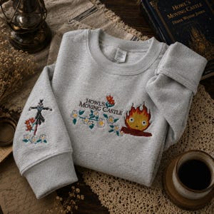 May include: A light gray sweatshirt with embroidered designs from "Howl's Moving Castle." The design includes the text "Howl's Moving Castle," a fire demon, and a scarecrow-like figure. Floral accents are also present. The sweatshirt is folded on a textured surface.