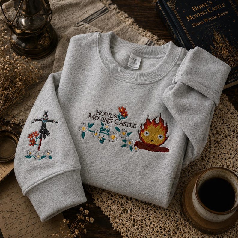 May include: A light gray sweatshirt with embroidered designs from "Howl's Moving Castle." The design includes the text "Howl's Moving Castle," a fire demon, and a scarecrow-like figure. Floral accents are also present. The sweatshirt is folded on a textured surface.