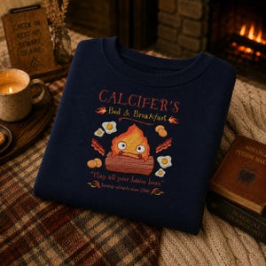 Puede incluir: Una sudadera azul marino de cuello redondo con un gráfico de un personaje de fuego de dibujos animados sentado sobre un tronco, rodeado de tocino y huevos. El texto dice: CALCIFER'S Bed & Breakfast, May all your bacon burn, Serving wizards since 1986.