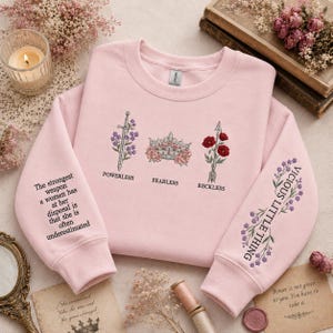Può includere: Una felpa girocollo rosa chiaro con ricami sul davanti e sulle maniche. Il davanti mostra una spada con fiori viola, una corona con rose rosa e una freccia con papaveri rossi, etichettati come Powerless, Fearless e Reckless.