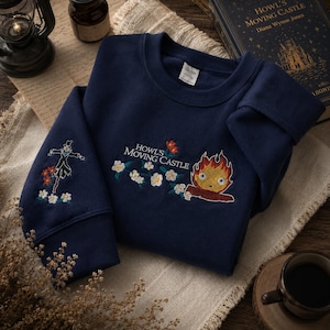 May include: Navy blue sweatshirt with embroidered designs from "Howl's Moving Castle." Features a scarecrow on one sleeve and a fire demon with the text "Howl's Moving Castle" on the chest, surrounded by flowers. The sweatshirt is folded on a neutral-colored cloth.